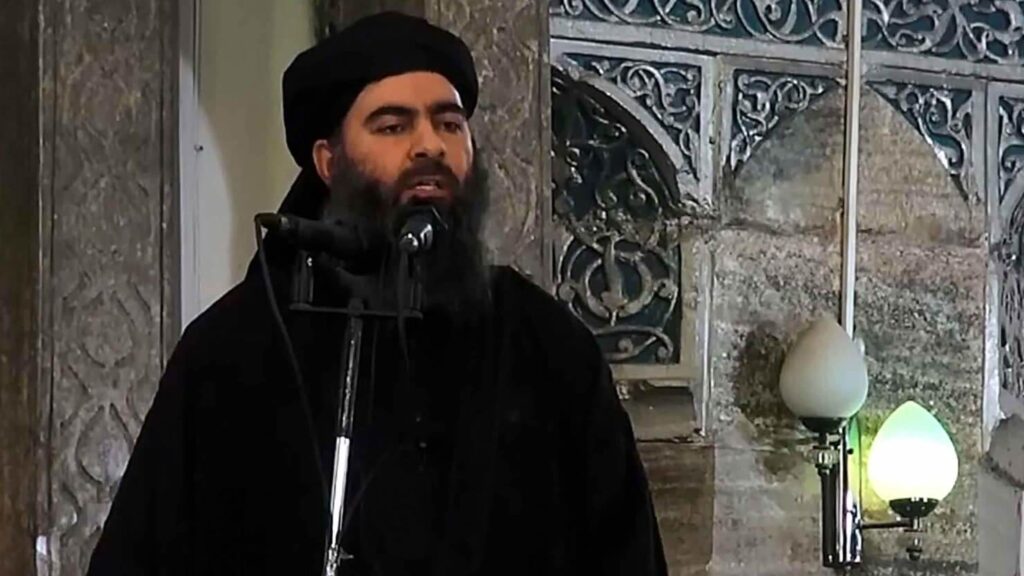 Man with a beard speaking into a microphone, wearing black clothing and a hat.