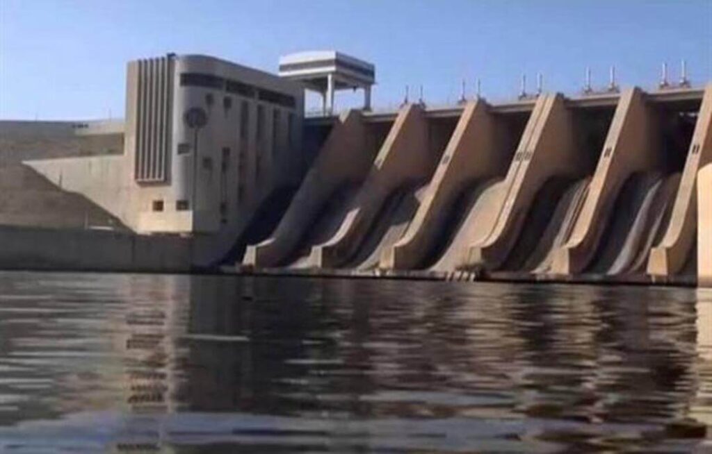 A large dam structure spanning a river under a clear blue sky.