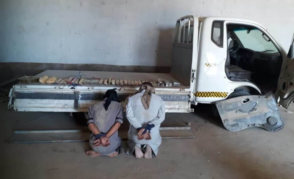 Two men sitting handcuffed beside a truck in a bare room.