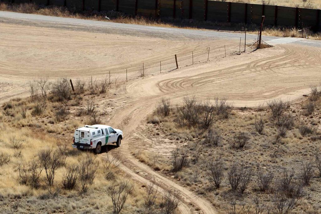 A white armored vehicle driving on a dusty dirt road in a barren landscape.