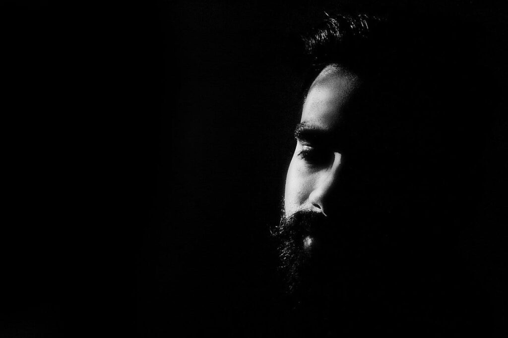 A dramatic black and white portrait of a bearded man in deep shadow.