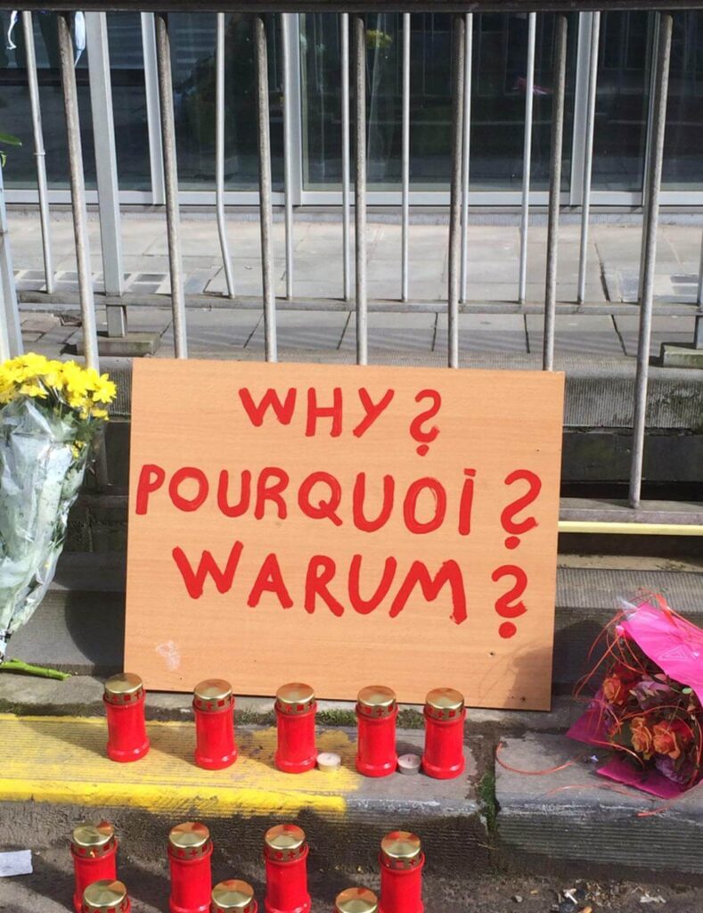 A cardboard sign asking 'Why?' in three languages with candles and flowers.