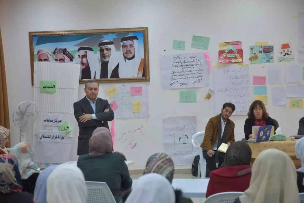 A man presenting to an audience in a room with posters and portraits of Middle Eastern leaders.