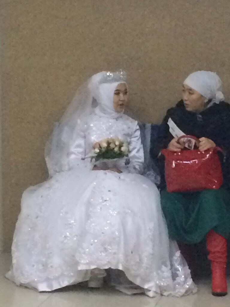 Bride in white gown sitting next to a woman with a red bag.