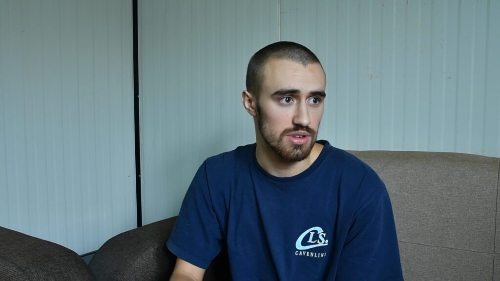 A man with a beard wearing a navy blue shirt sitting on a couch indoors.