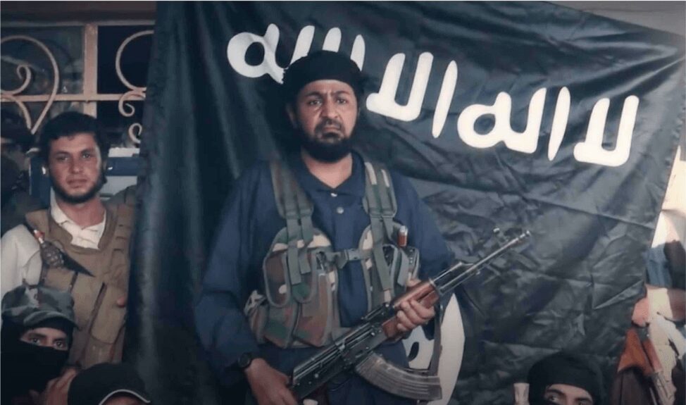 Man with rifle and tactical vest stands in front of black flag with Arabic script.
