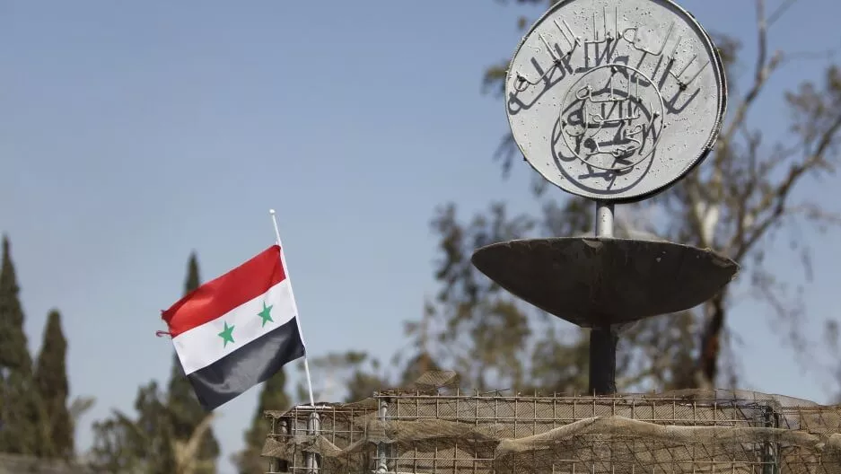 Syrian flag waving near a memorial with Arabic inscription.