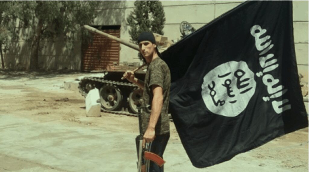 A militant holding a gun and a black flag with Arabic script, standing near a military vehicle.