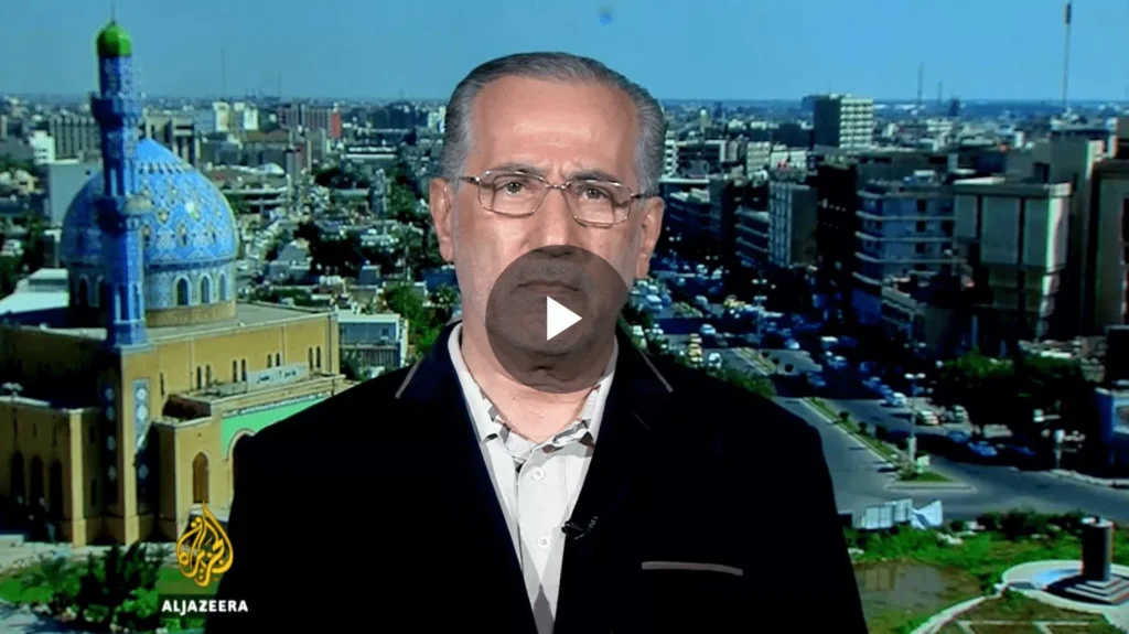 A man with glasses speaking on camera in a city backdrop.