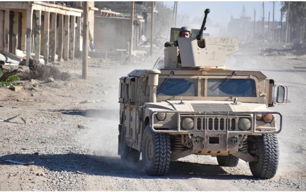 Military Humvee driving through a dusty urban area with a mounted weapon.