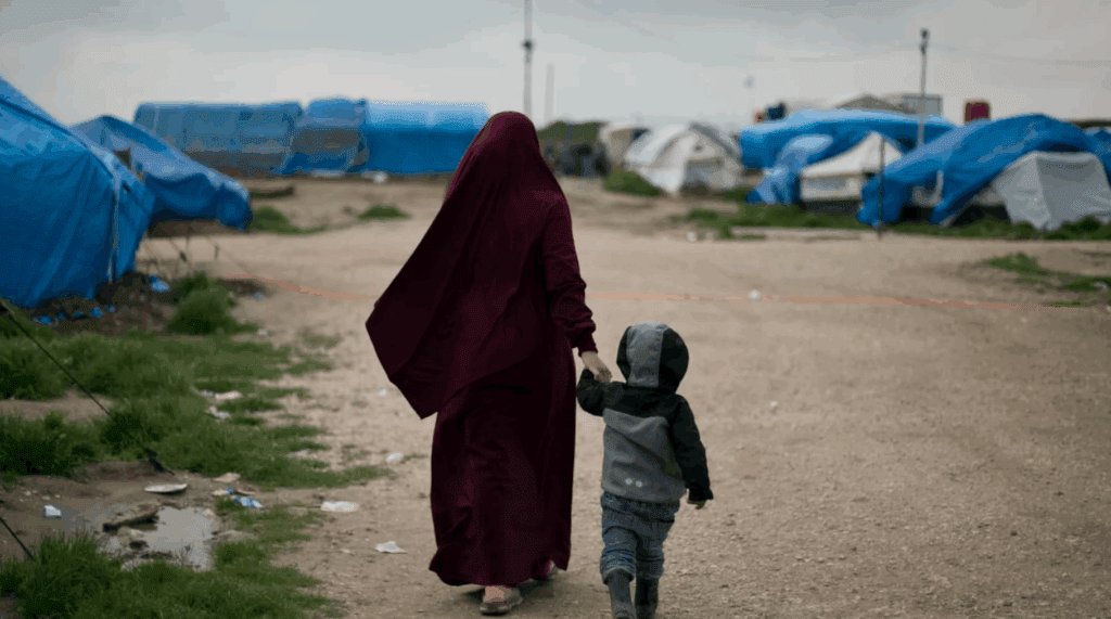 A woman in a maroon cloak walking hand-in-hand with a child in a refugee camp.