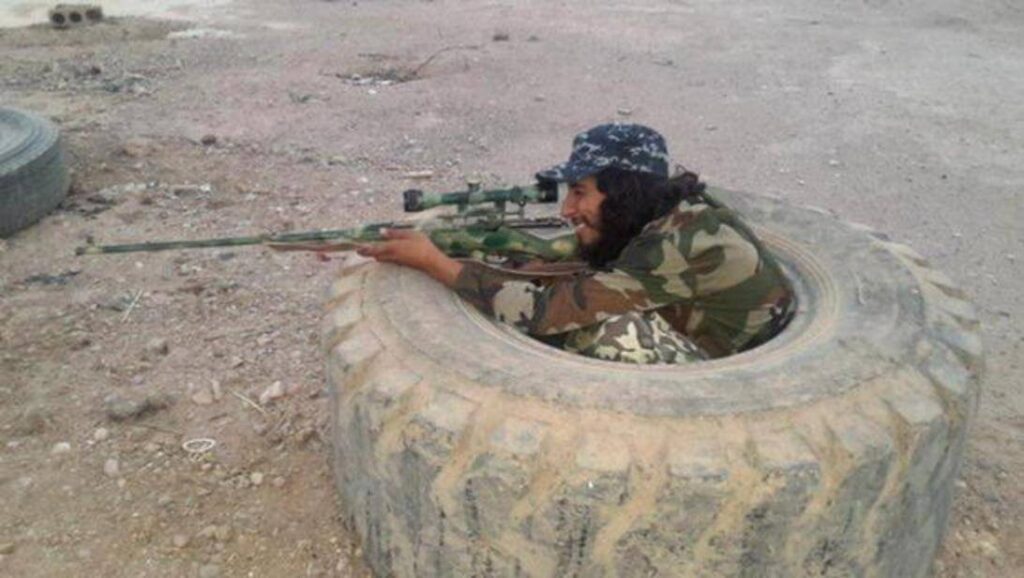 Person aiming a rifle from behind a tire in a desert area.