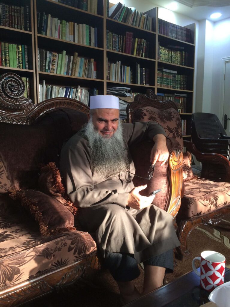 An elderly man with a long white beard sitting on an ornate sofa in a cozy library.