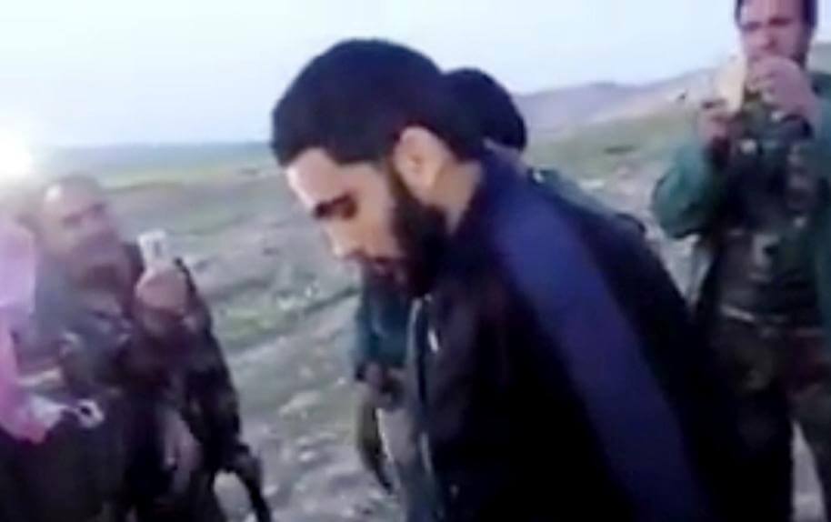 A man with a beard and tied-back hair stands outdoors with others.