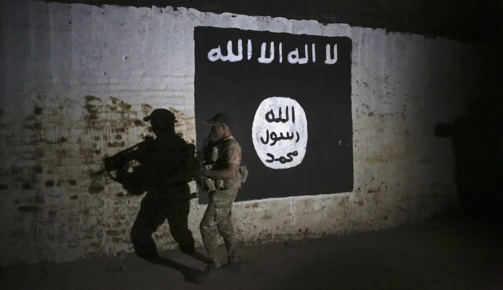 Two armed individuals stand near a black flag with white Arabic text on a wall.