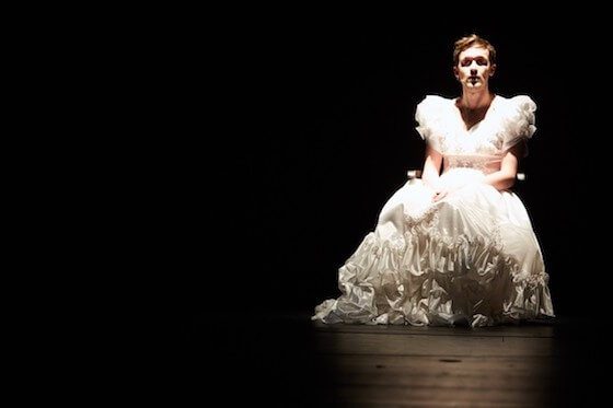 A woman in a white dress sitting alone on a dark stage.