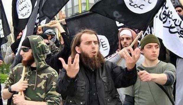 A group of men in militant attire raising their hands with flags in the background.