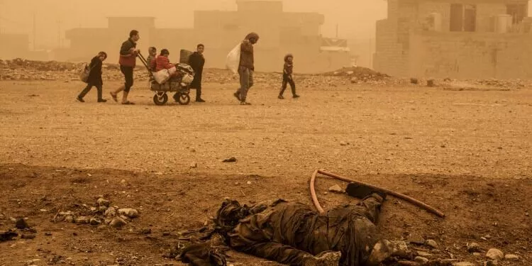 People walk through a dusty street with debris and a dead body in the foreground.