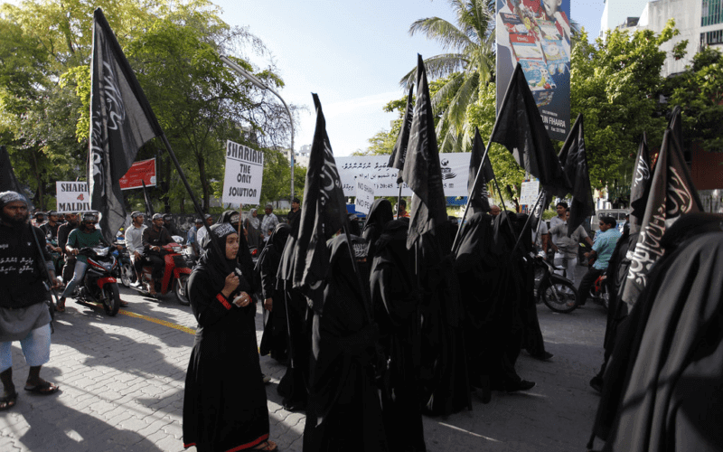 Women dressed in black robes holding black flags during a public gathering.