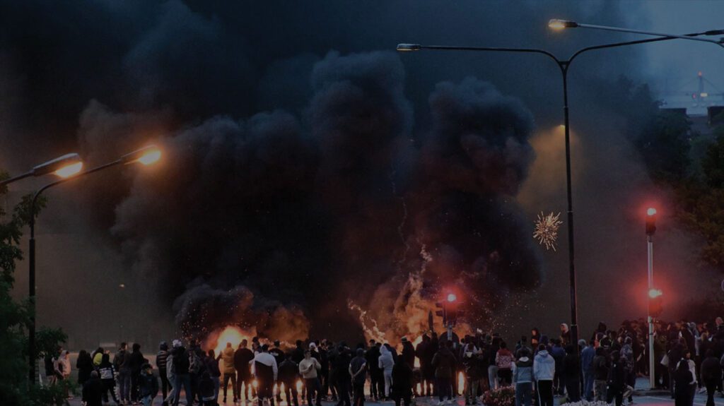 Crowd watches large fire emitting thick black smoke at night.