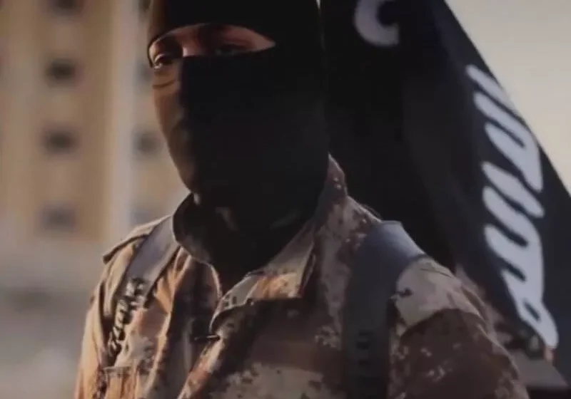 A masked militant in camouflage stands before a black flag with Arabic script.