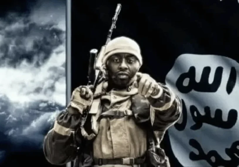 Soldier in combat gear pointing forward with a flag in the background.