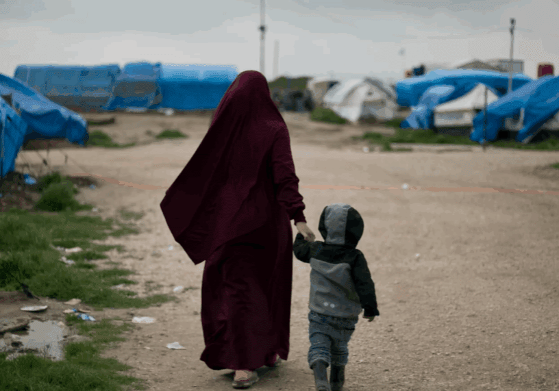 A woman in a maroon cloak walking hand-in-hand with a child in a refugee camp.