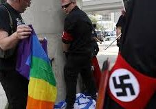A man in black with a Nazi armband stands near a rainbow flag and a Nazi flag.