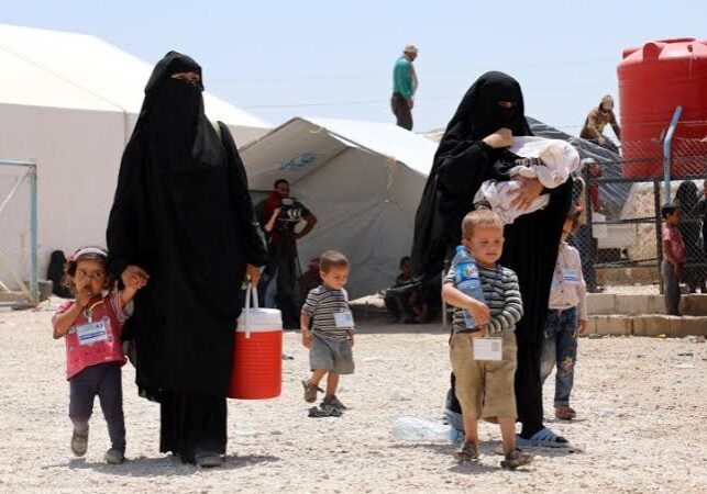 Women in black abayas and children at a refugee camp tent.